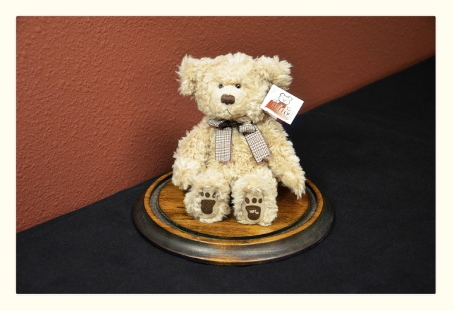 A plush teddy bear with a checkered bow tie sits on a round wooden base against a red wall, offering comfort akin to that sought by ketamine therapy patients. The bear's paws feature paw print designs, and a small tag is attached to its ear. The base rests on a dark surface. - Ketamine